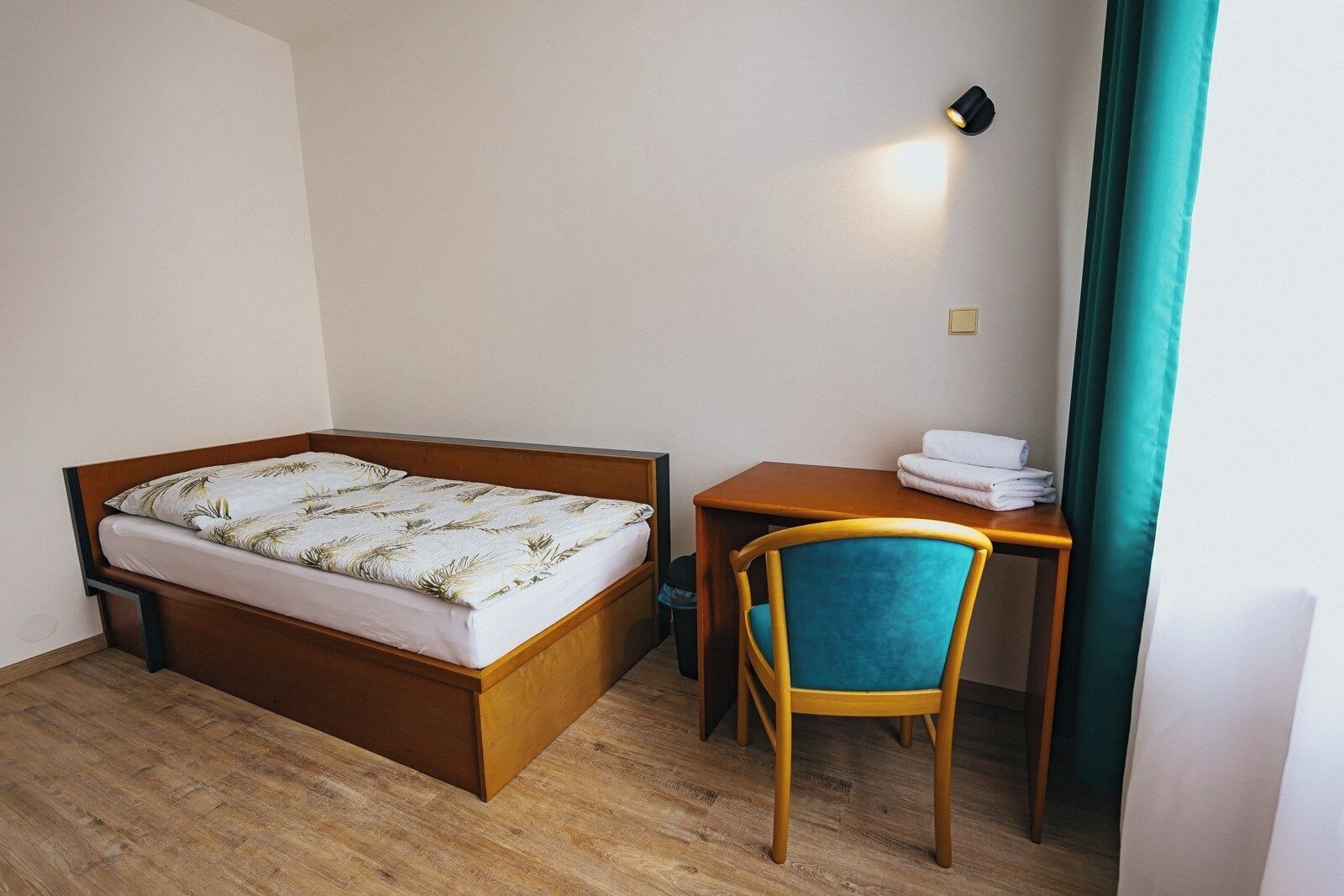 A single bed with a wooden frame, a small desk with a chair, and a wall-mounted light.