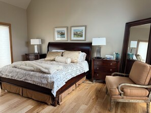 A bedroom with a large bed, wooden floor, two bedside tables with lamps, a chair, and two framed pictures on the wall.