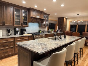 A modern kitchen with dark wood cabinets, granite countertops, a large island with bar stools, and a dining area with leather chairs.