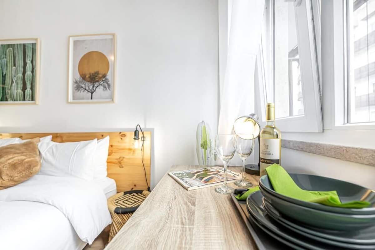 A hotel room with a wooden headboard, a bed with white bedding, a wooden table with a magazine, two wine glasses, a bottle of wine, and a green bowl.