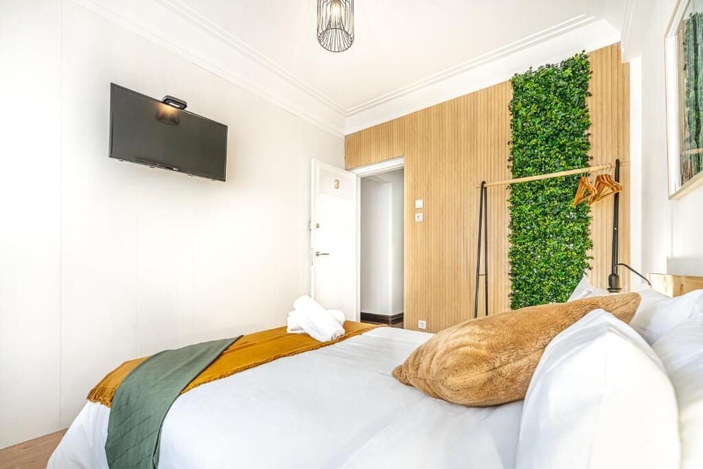 A hotel room with a bed, a mounted TV, a green vertical garden wall, and a wooden door.