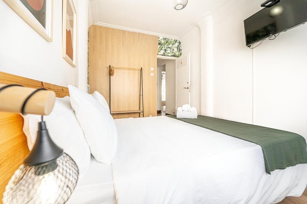 A neatly made bed with white bedding and a green throw, a wooden headboard, a wall-mounted TV, and a door leading to another room.