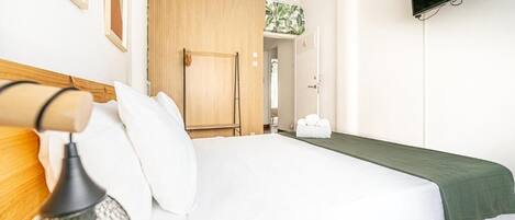 A neatly made bed with white bedding and a green throw, a wooden headboard, a wall-mounted TV, and a door leading to another room.