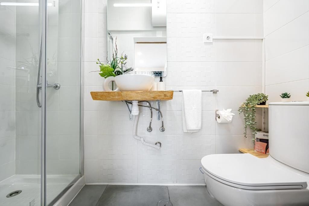 Un baño moderno con cabina de ducha de vidrio, inodoro blanco y estante de madera con lavamanos blanco y plantas.