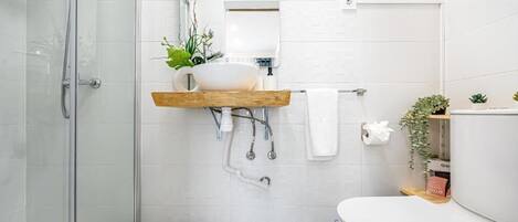 Un baño moderno con cabina de ducha de vidrio, inodoro blanco y estante de madera con lavamanos blanco y plantas.
