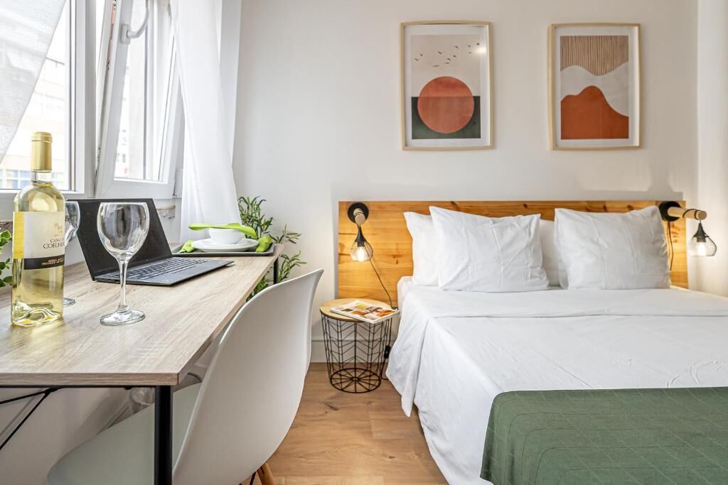 Une chambre d’hôtel avec une tête de lit en bois, du linge de lit blanc, un bureau avec un ordinateur portable et une bouteille de vin, ainsi que deux tableaux abstraits accrochés au mur.