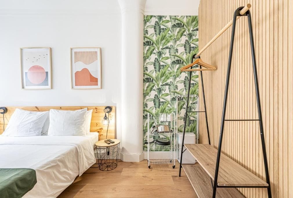 Une chambre moderne avec une tête de lit en bois, un lit recouvert de linge de maison blanc, une petite table ronde, un luminaire suspendu et un mur recouvert d’un papier peint à motifs tropicaux.