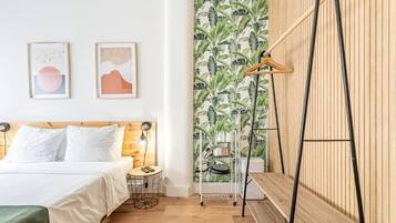 Une chambre moderne avec une tête de lit en bois, un lit recouvert de linge de maison blanc, une petite table ronde, un luminaire suspendu et un mur recouvert d’un papier peint à motifs tropicaux.