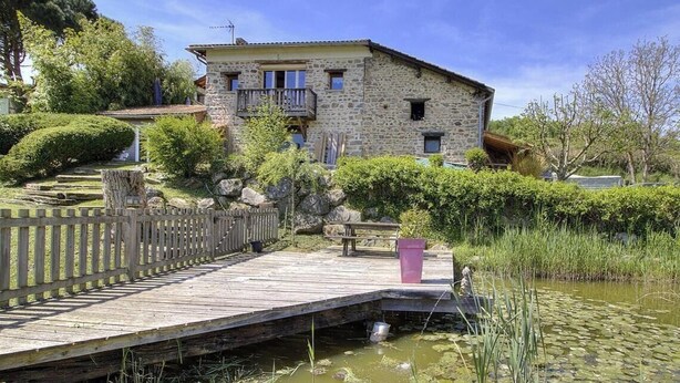 Un edificio de piedra con un balcón, una terraza de madera y un estanque con nenúfares.