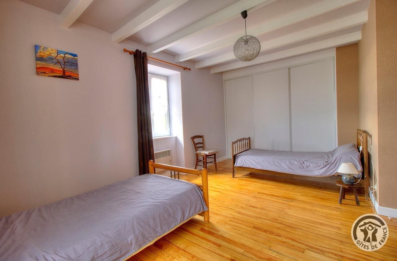 Een kamer met twee eenpersoonsbedden, een houten vloer, een raam met gordijnen, een stoel en een hanglamp.
