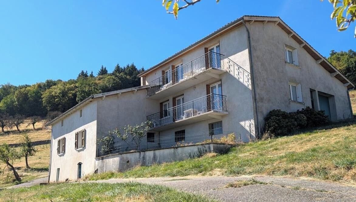 A two-story building with a balcony on the upper floor, surrounded by a grassy landscape.