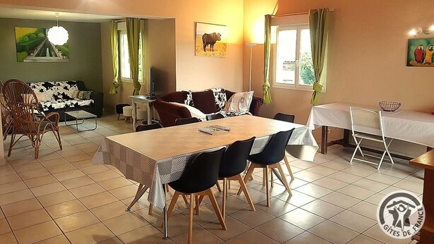 Ein Essbereich mit großem Tisch, Stühlen und einer Couch. An der Wand hängt ein Gemälde und das Fenster ist mit Vorhängen versehen.