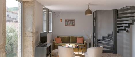 Un salon moderne avec un canapé, deux fauteuils, une table basse et un téléviseur. Un escalier se trouve à droite et une grande fenêtre à gauche.