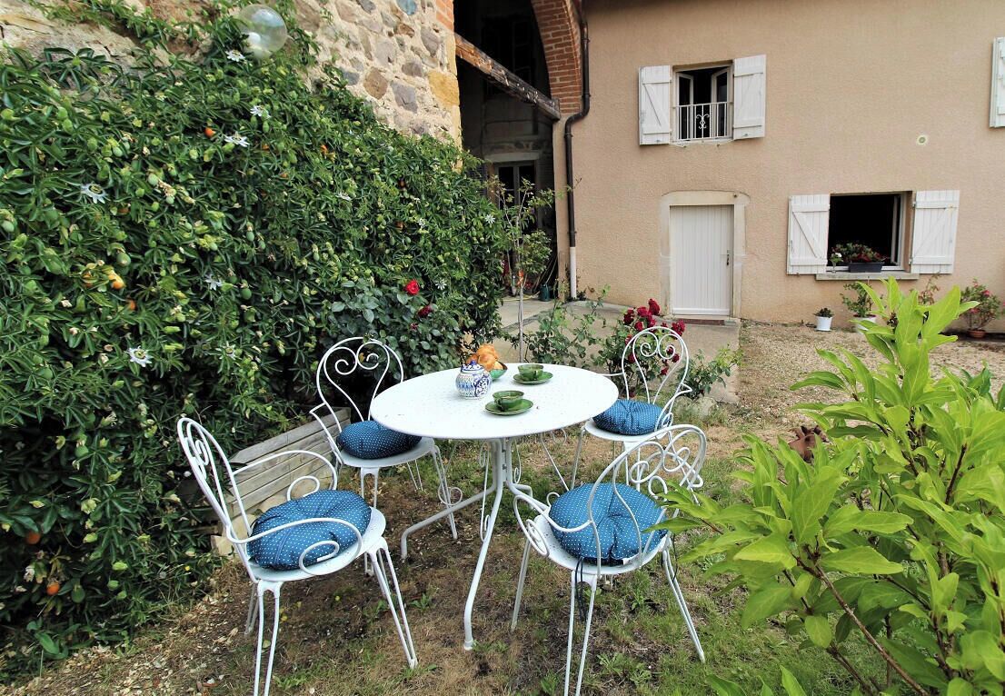 Un jardin avec une table ronde et des chaises en métal blanc, entouré de verdure et bordé d’un bâtiment aux volets blancs.