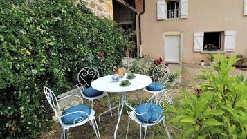 Un jardin avec une table ronde et des chaises en métal blanc, entouré de verdure et bordé d’un bâtiment aux volets blancs.