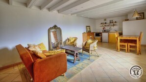 Una sala de estar con un sofá, sillones, una mesa de centro y una mesa de comedor con sillas. Hay una zona de cocina con un fregadero, estufa y estanterías con utensilios de cocina.