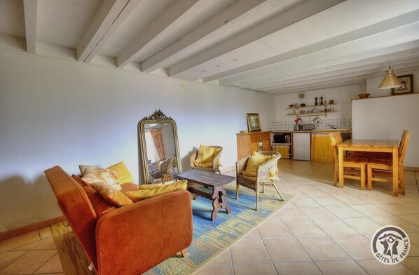 A living room with a sofa, armchairs, a coffee table, and a dining table with chairs. There is a kitchen area with a sink, stove, and shelves with kitchenware.