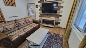 A living room with a sofa, a coffee table, a TV mounted on a wooden wall, and a window with curtains. - ENTIRE LAKEFRONT 10-bedroom Chateau Getaway at Whitecap Mountains Resort (Upson)