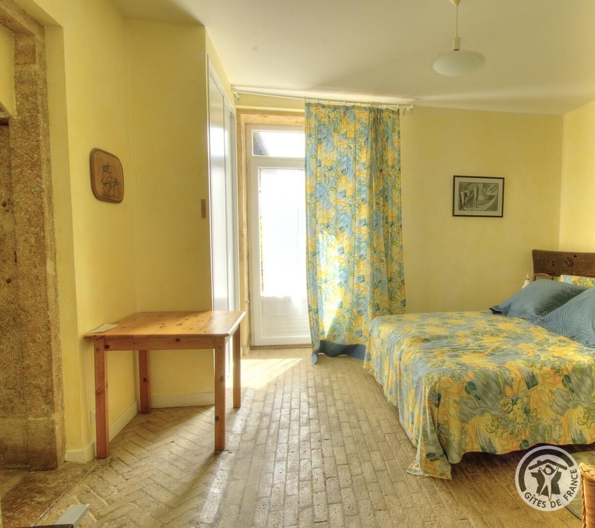 Ein Zimmer mit einem Bett, einem Holztisch, einem Fenster mit Vorhängen und einem Bild an der Wand.
