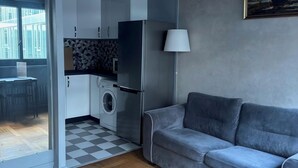 A modern living room with a grey sofa, a wooden herringbone floor, a kitchen with white cabinets, a washing machine, and a painting on the wall. - Central Cozy Apartment Paris 2nd District (Paris)