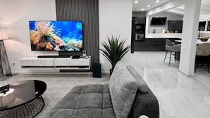 A modern living room with a large flat-screen TV, a grey sofa, and a dining area in the background.