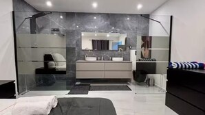 A modern bedroom with a large bed, a glass shower enclosure, a vanity with a mirror, and a dark-colored rug.