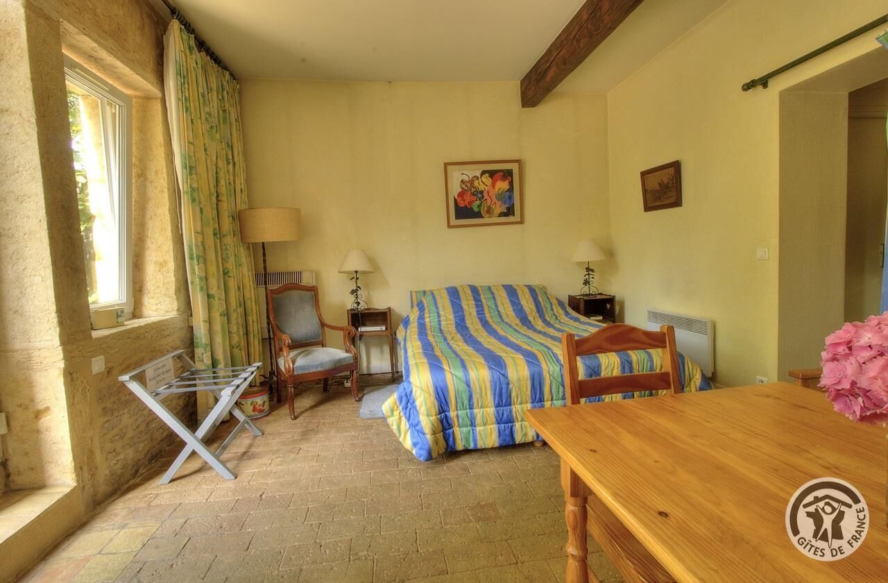 A room with a bed, a wooden table, a chair, a lamp, and a window with curtains.
