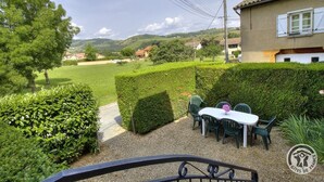 Un patio con mesa y sillas, rodeado de setos bien cuidados y vistas a un paisaje rural con colinas y casas.