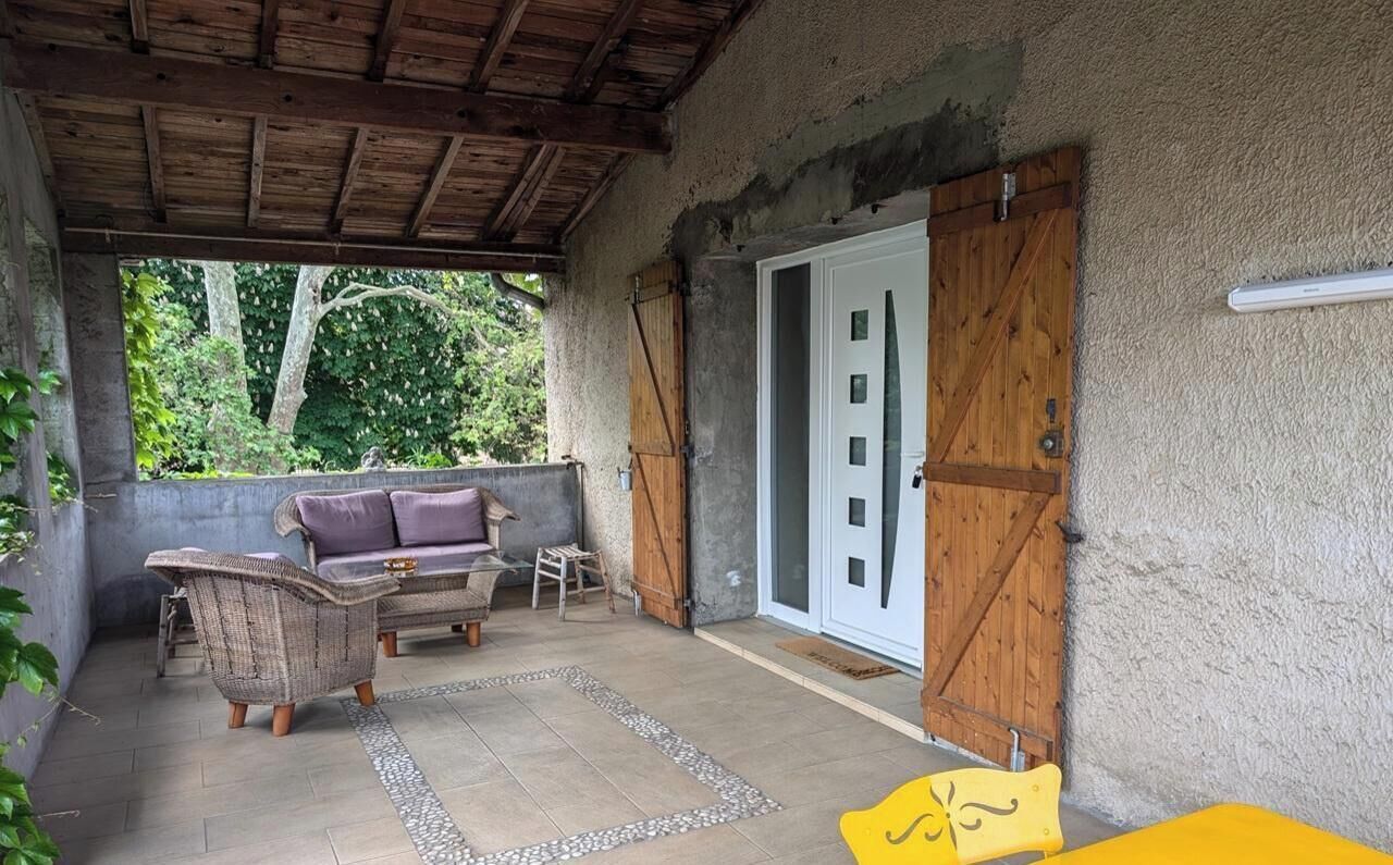 Une terrasse couverte avec un canapé et des fauteuils, une porte en bois et un mur en béton.