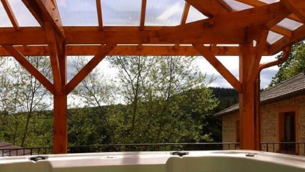 Een jacuzzi onder een houten pergola, met uitzicht op bomen en een gebouw op de achtergrond.