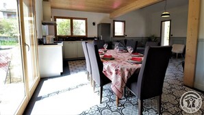 Uma sala de jantar com mesa posta para quatro pessoas, cozinha ao fundo e vista para o exterior através de grandes janelas.