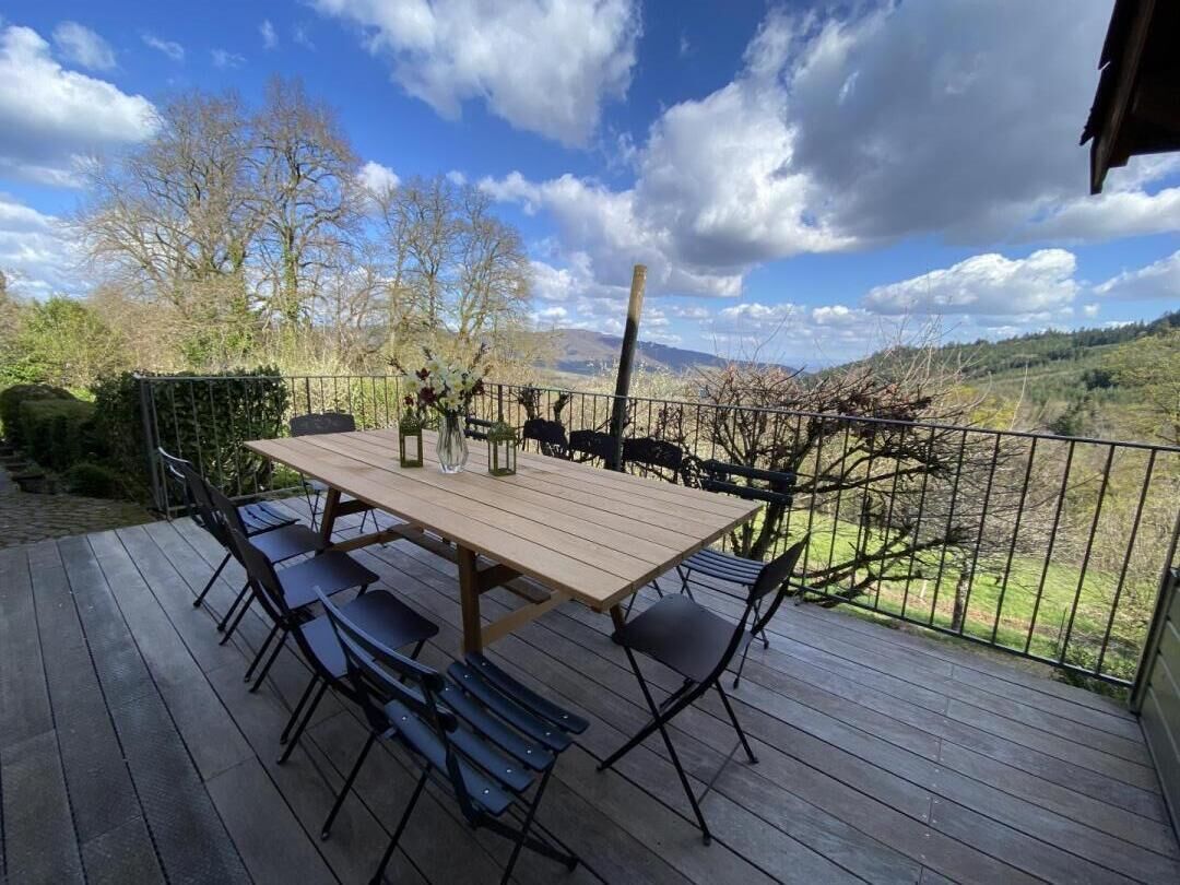 A wooden deck with a table and chairs, surrounded by a metal fence, overlooking a hilly landscape.