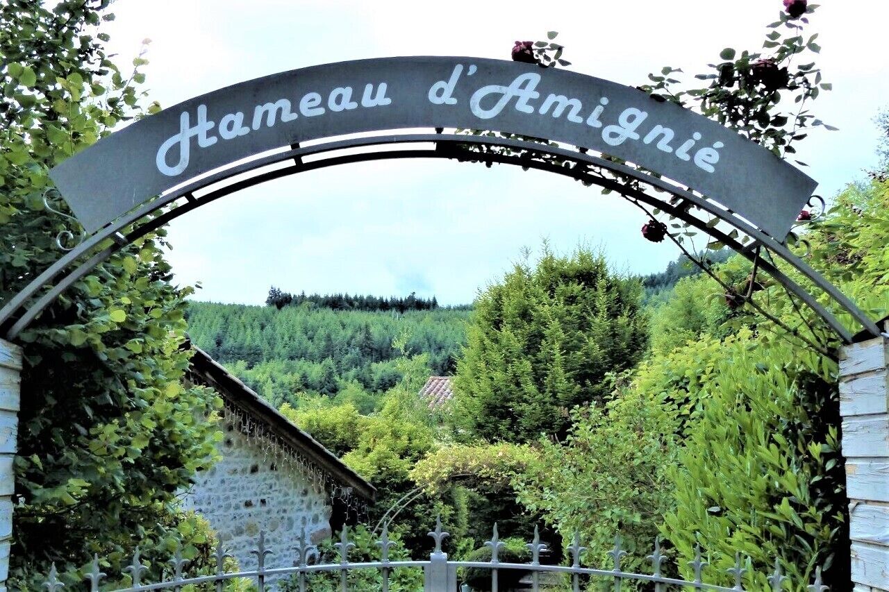 A metal archway with the inscription "Hameau d'Amigny" in cursive, leading to a lush, green landscape with trees and a stone building in the background.