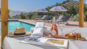 Escena junto a la piscina con sillones de descanso, una botella de vino Penascal, un sombrero de paja, una toalla y un libro sobre una mesa.
