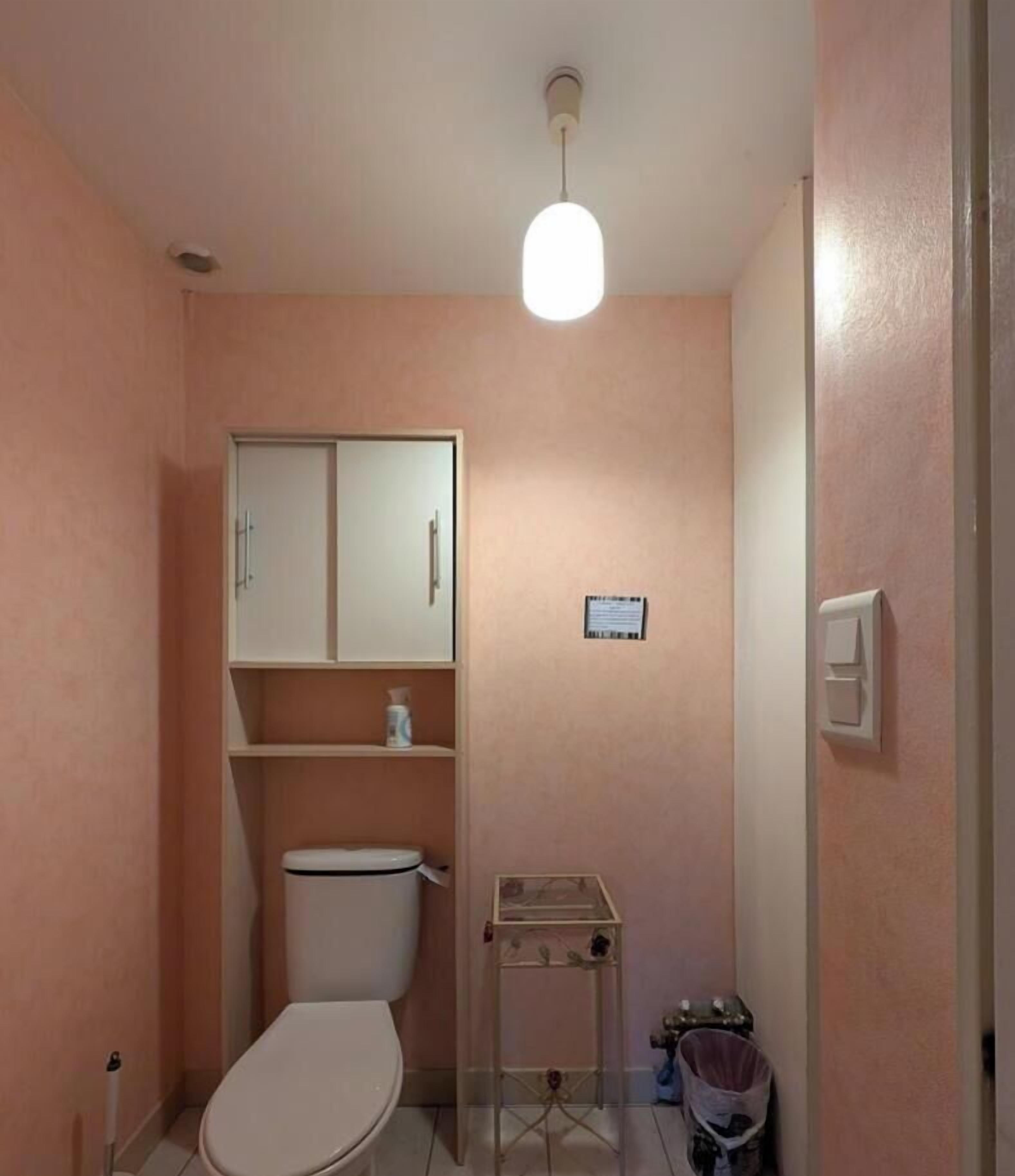 A small bathroom with a toilet, a cabinet, a shelf with toiletries, a small table, and a hanging light fixture.