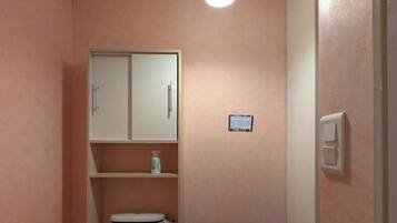 A small bathroom with a toilet, a cabinet, a shelf with toiletries, a small table, and a hanging light fixture.