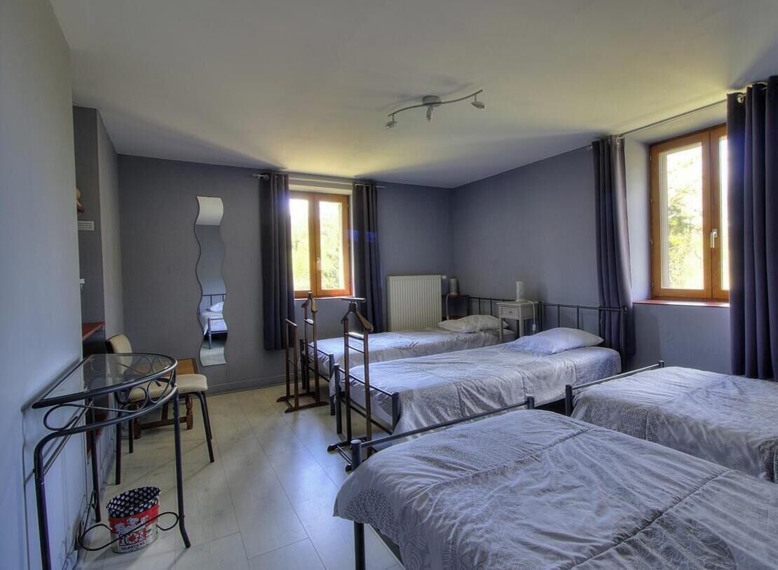 A room with three beds, a small table, a chair, a radiator, and a window with curtains.