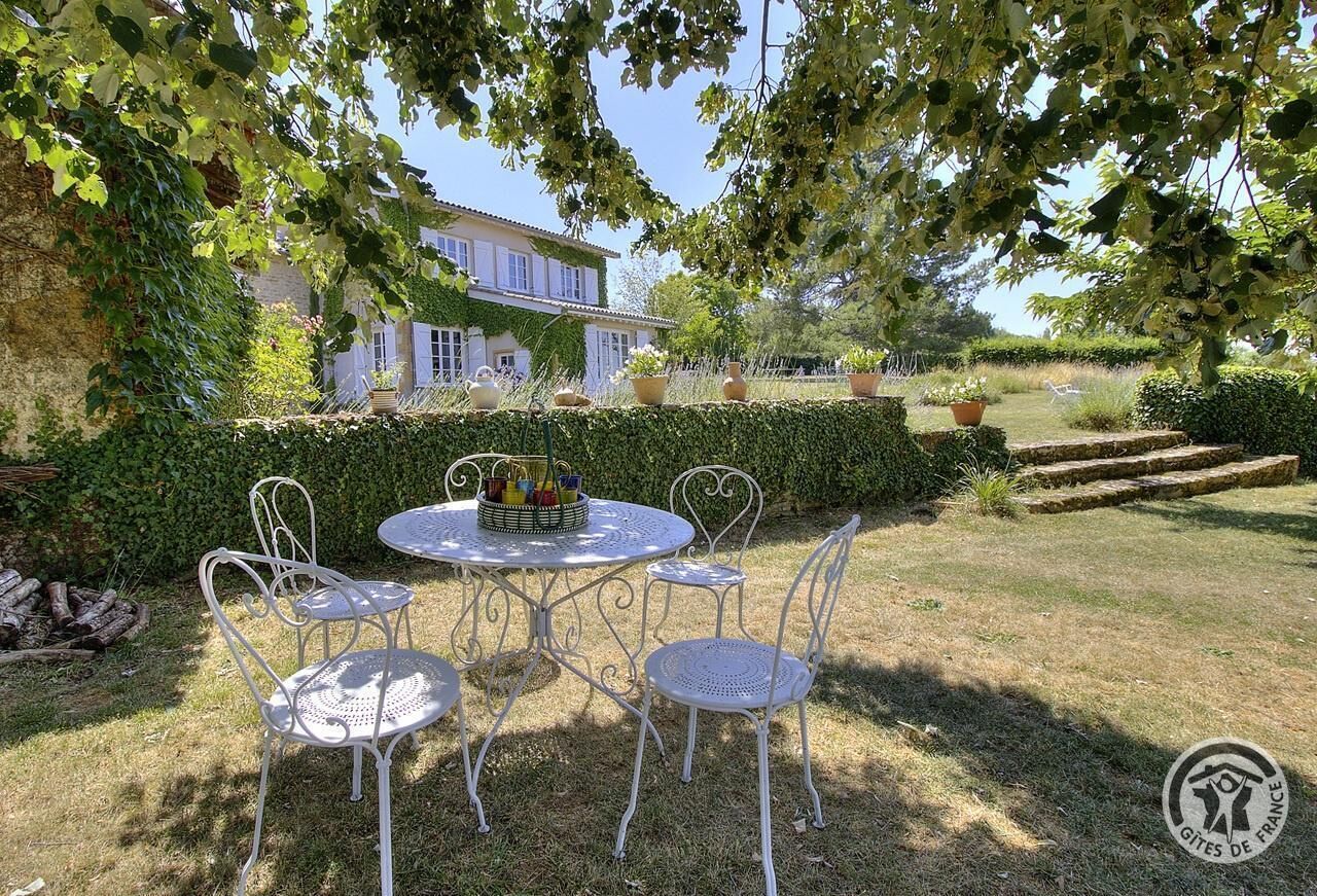 Un espace extérieur aménagé avec une table ronde et quatre chaises, entouré d’une haie bien entretenue et avec une maison en arrière-plan.