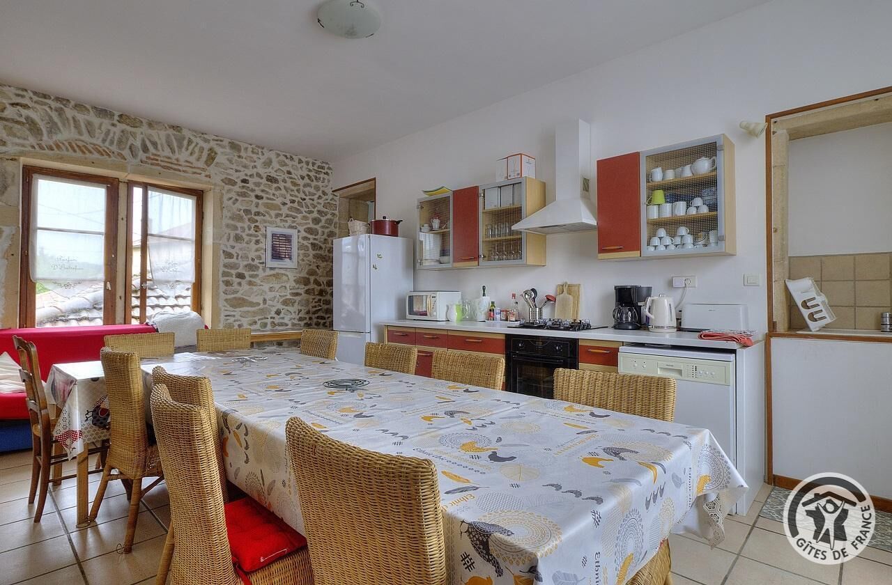 A dining area with a long table and wicker chairs, a kitchen with a stove and cabinets, and a stone wall.