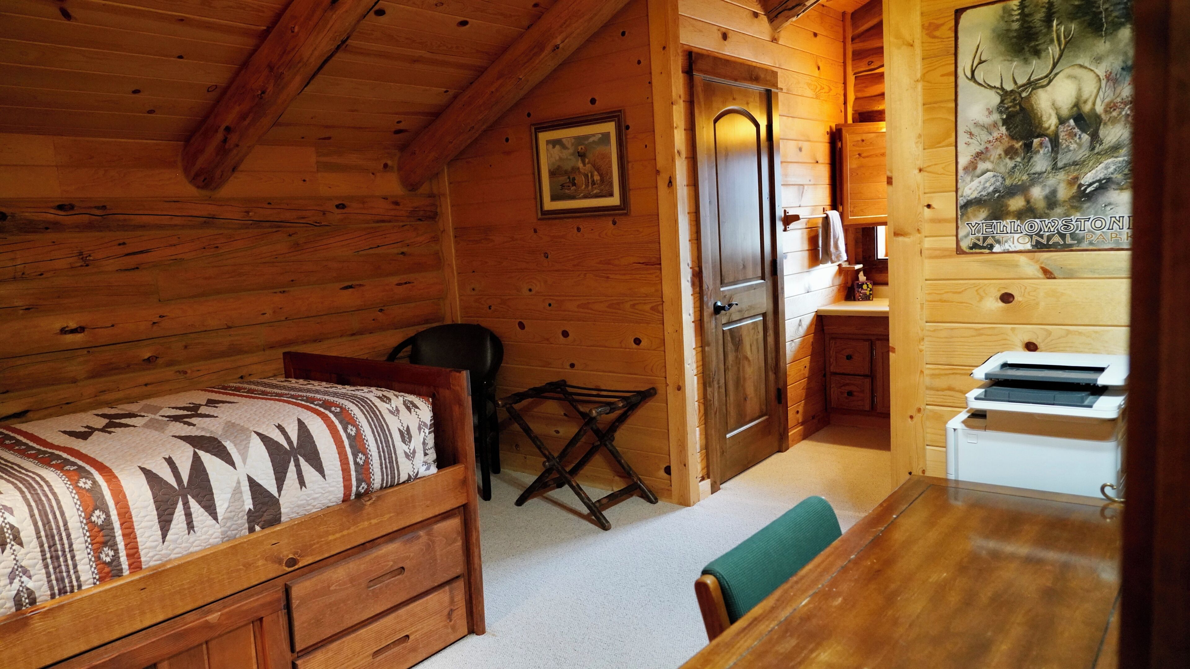 L’intérieur d’une cabane en bois, avec un lit, un bureau, une chaise et une imprimante.