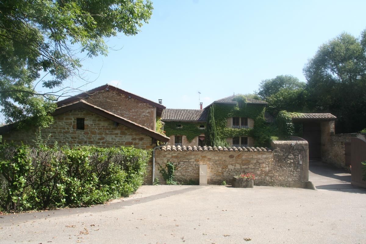 Un bâtiment en pierre avec un toit recouvert de tuiles, une allée pavée et un petit jardin agrémenté d’arbustes et d’une poubelle.
