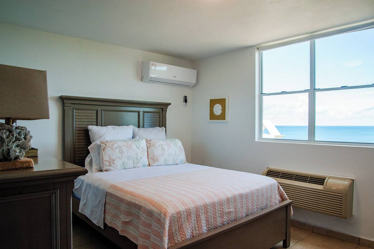 A bedroom with a bed, a nightstand, an air conditioning unit, a window with a view, and a wall-mounted heater.