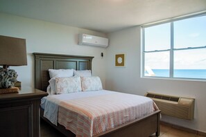 A bedroom with a bed, a nightstand, an air conditioning unit, a window with a view, and a wall-mounted heater. - El Combate Oceanview Condos (Boquerón)