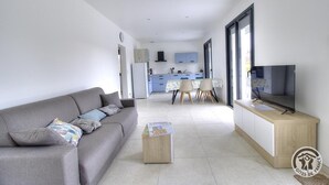 Una sala de estar moderna con un sofá gris, una mesa de centro y un televisor sobre un mueble de madera. Se ve una cocina con armarios azules y una zona de comedor con una mesa y sillas al fondo.
