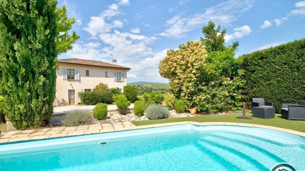 Une piscine à l’eau d’un bleu limpide, entourée d’un jardin bien entretenu où poussent différentes espèces d’arbustes et un grand arbre.