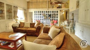 Un salón espacioso con un conjunto de sofá de cuero marrón, una mesa de centro de madera y un sillón blanco. Un aparador de vidrio con objetos decorativos se encuentra a la izquierda, y una escalera conduce a un nivel superior.