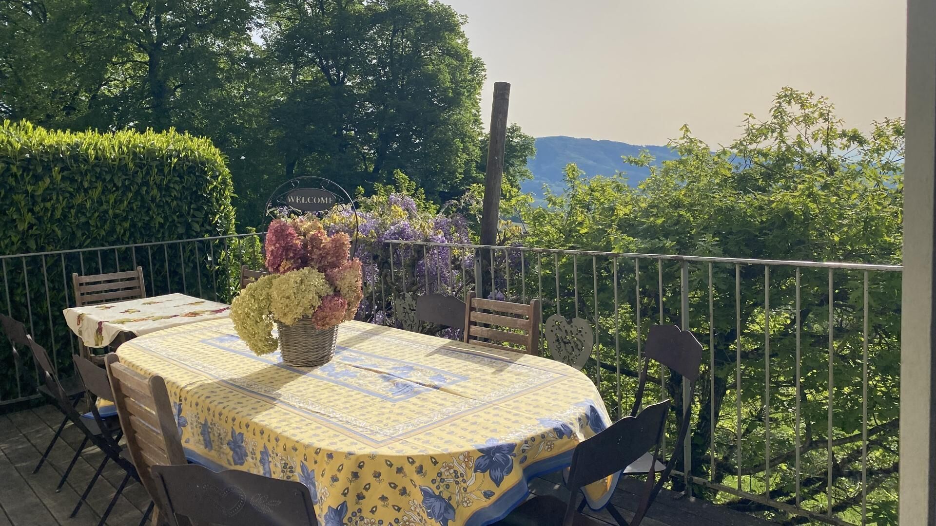 Ein Tisch im Freien mit floralem Mittelstück, umgeben von Stühlen, auf einem Balkon mit Blick auf Bäume und Berge.