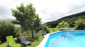 Une piscine dotée d’une terrasse blanche, entourée de chaises vertes et d’un arbre.