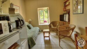 A compact kitchen with a microwave, coffee maker, and utensils. A bed with a green blanket is against the wall, and a window provides a view of greenery. Two wicker chairs are near a small table.