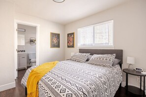 Ein Schlafzimmer mit einem Bett, einem Nachttisch mit Lampe, einem Fenster mit Jalousien und einem durch eine offene Tür einsehbaren Badezimmer.